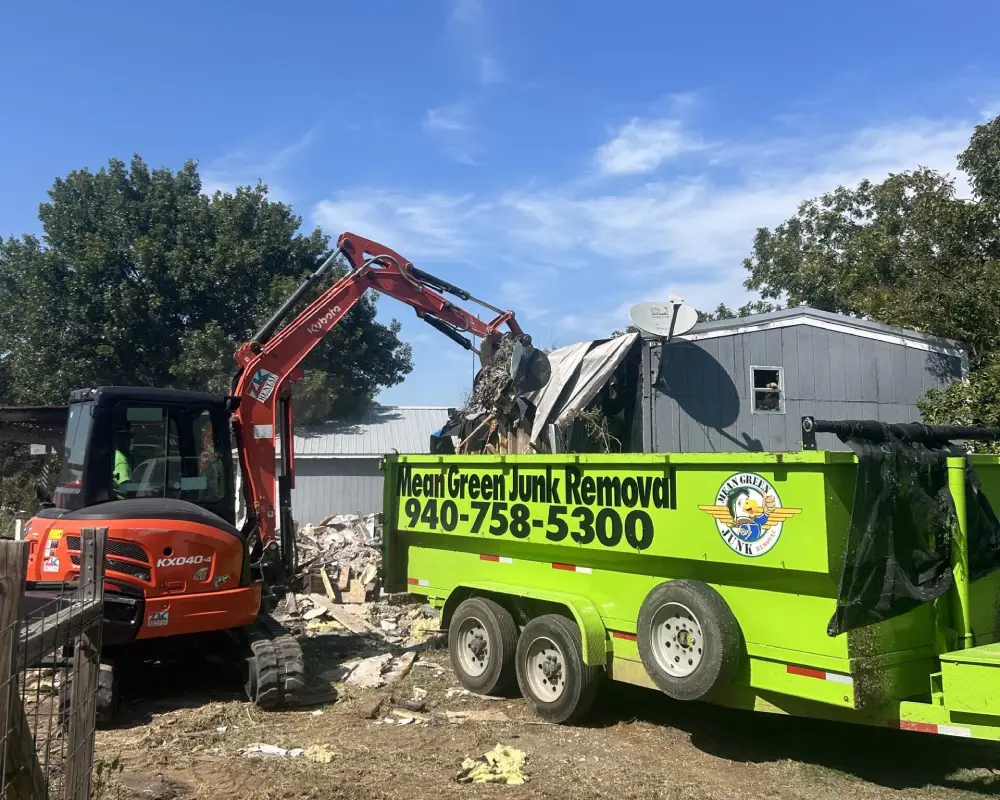 Excavator loading demolition debris into a Mean Green Junk Removal trailer at a cleanup site.
