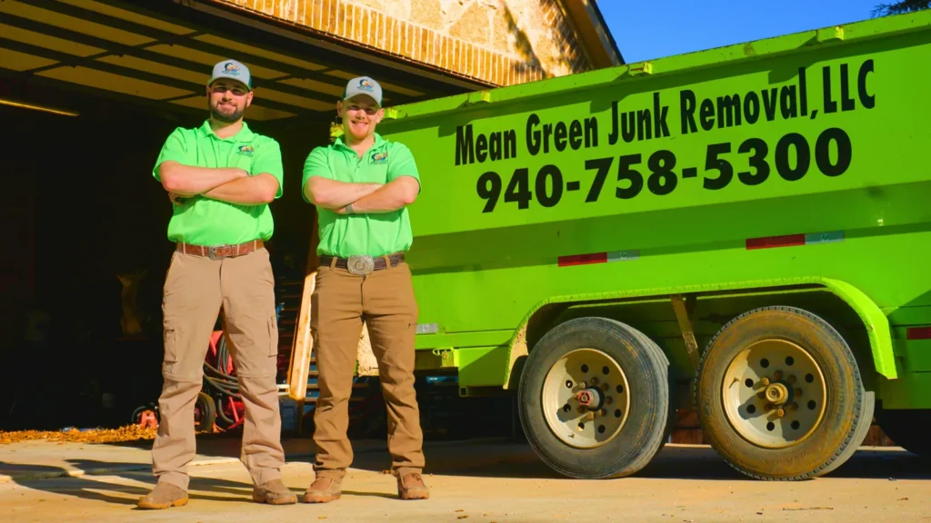Two Mean Green Junk Removal team members standing beside the branded trailer showing the company name and phone number 940-758-5300