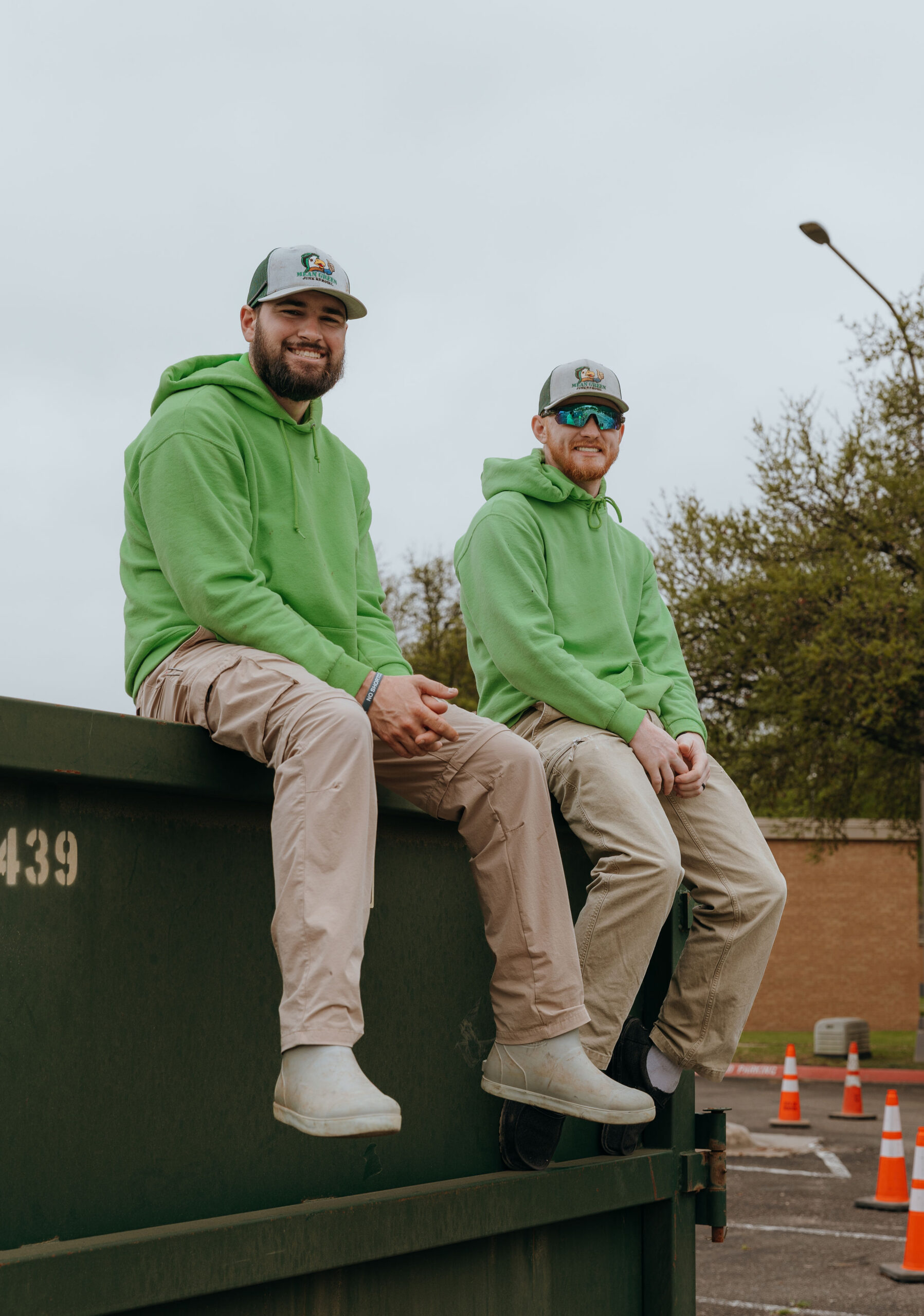 Two Mean Green Junk Removal crew members sitting on a dumpster during a cleanup service.